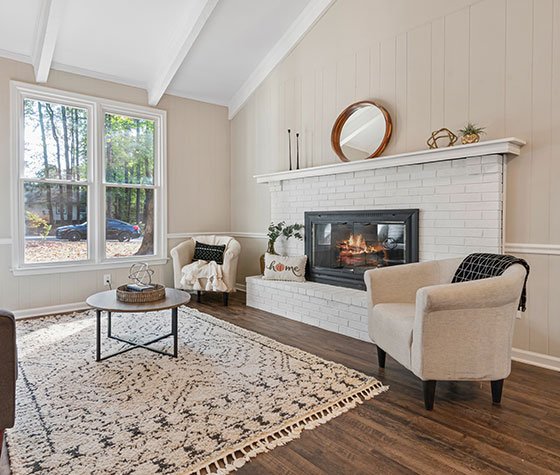 A spacious section of the living room featuring a large fireplace with white brick surround, cream siding on the walls, and a dark wooden floor. Two elegant white cream armchairs complement the cozy ambiance, while a big window floods the space with natural light, creating a warm and inviting atmosphere.