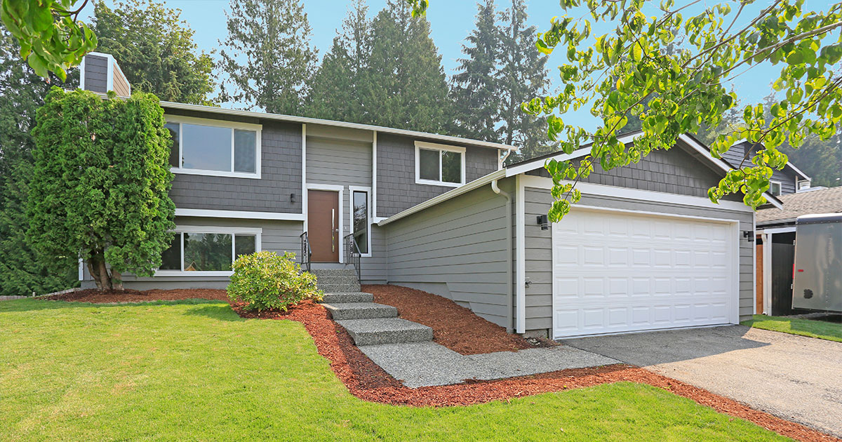 Modern suburban home exterior with manicured lawn, gray siding, white garage door, and lush green landscaping