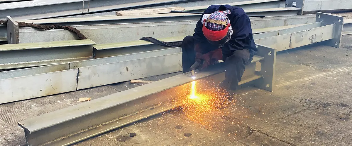 Worker welding steel beams for construction in an industrial setting, showcasing metal fabrication techniques.