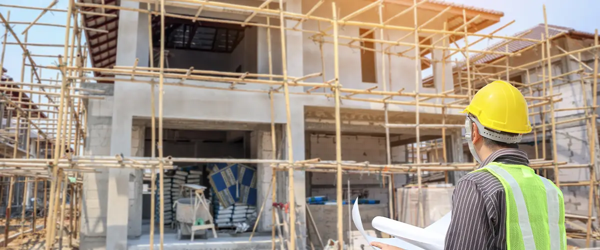 Construction worker in a yellow hard hat inspecting a residential building under construction with bamboo scaffolding.