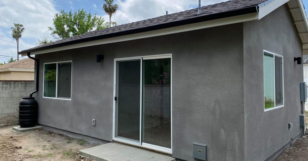 Newly built ADU with gray stucco exterior, sliding glass door, and rainwater barrel in a California backyard