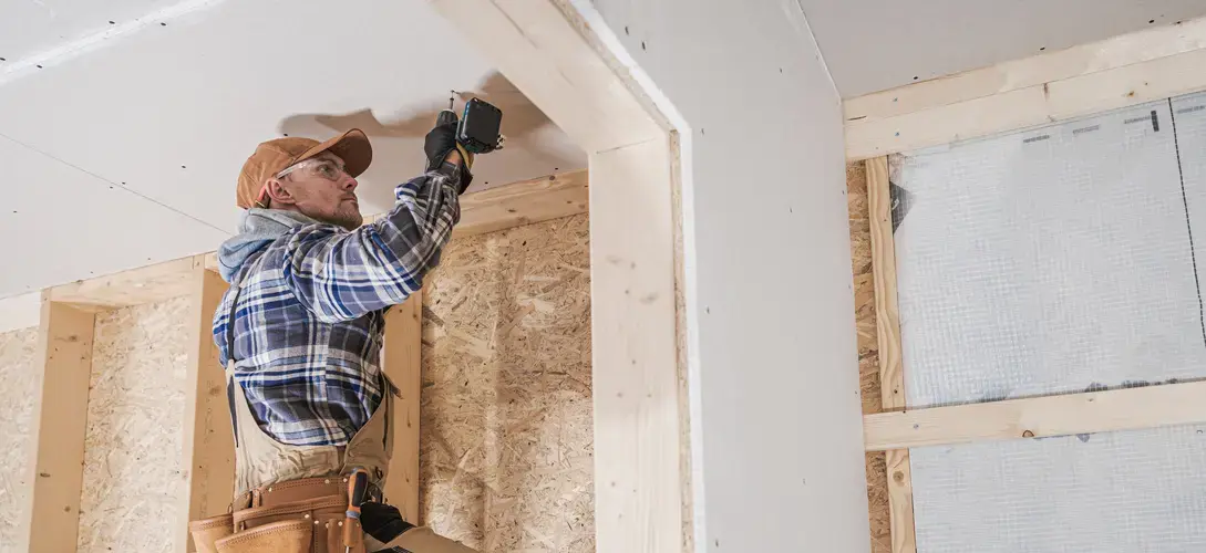 Construction worker installing ceiling drywall in a framed interior space, using a power drill while standing on a ladder