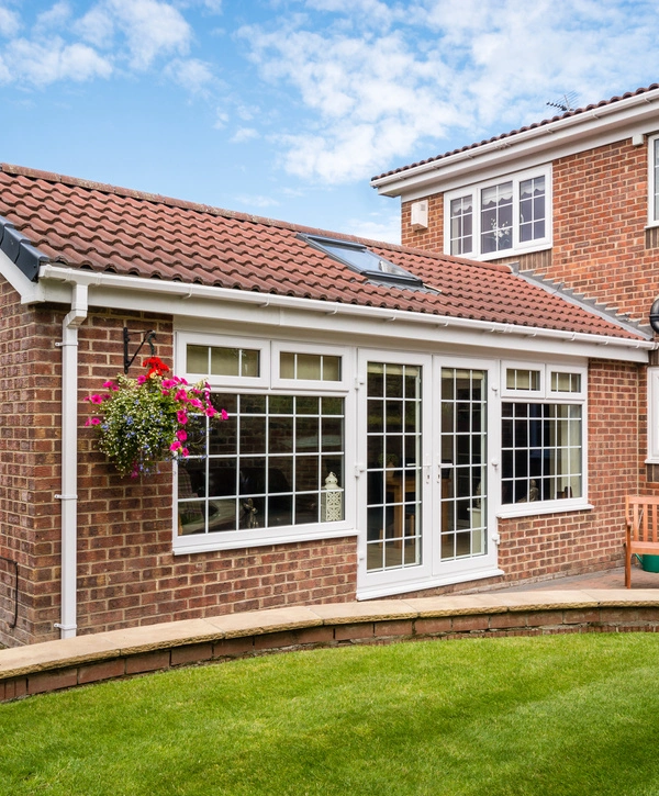 A sunny view of a red brick house extension (like many modern ADU homes) with a terracotta tile roof, white UPVC French doors, a large grid window, and a hanging flower basket overlooking a lush green lawn.