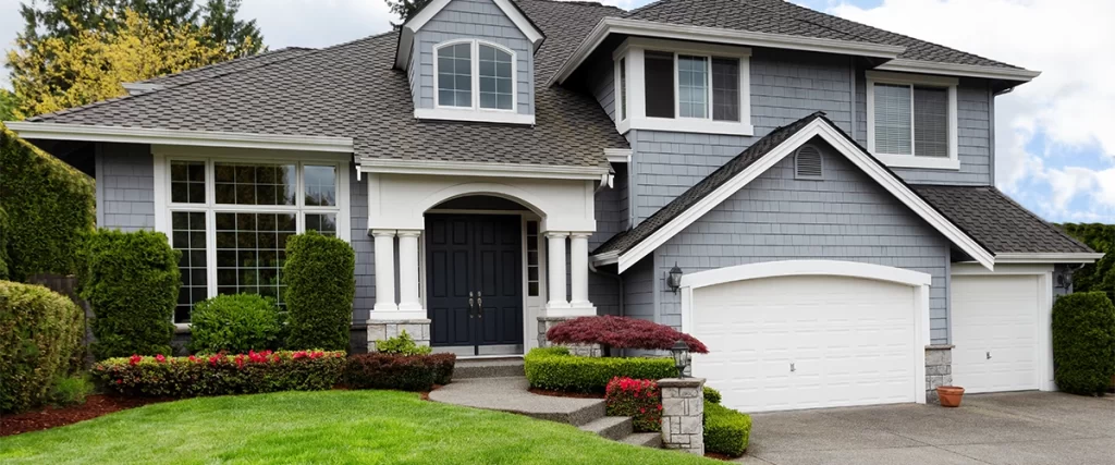 A traditional two-story residential home with gray shingle siding, white trim, columns at the entryway, and two white garage doors, surrounded by well-manicured hedges and lawn.