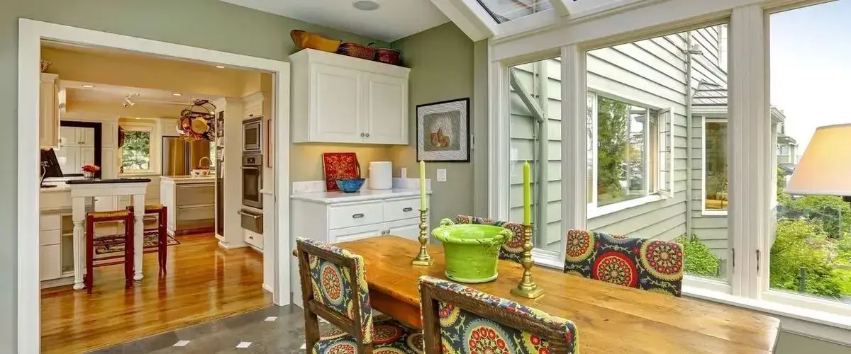 A sunlit craftsman-style dining room featuring a long wooden table, colorful patterned chairs, and a view into a modern white kitchen with hardwood floors.