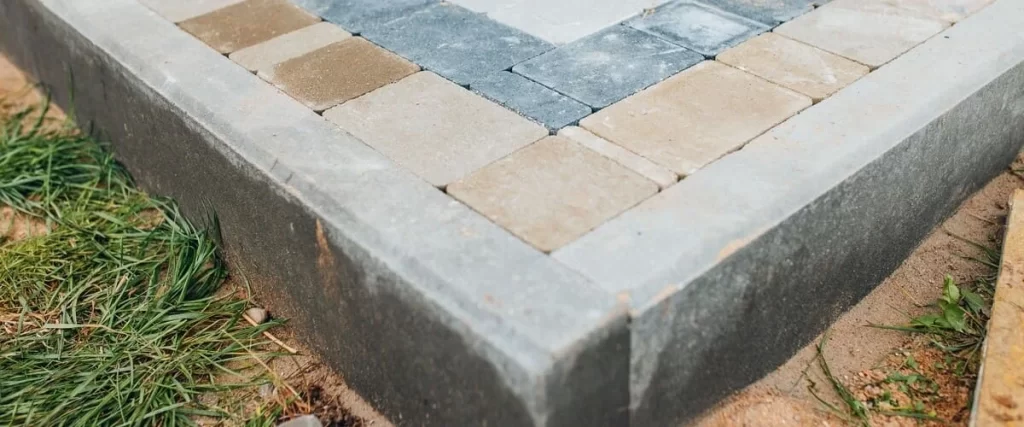 A close-up view of tan and grey interlocking paving stones neatly installed against a solid concrete building foundation corner outdoors.