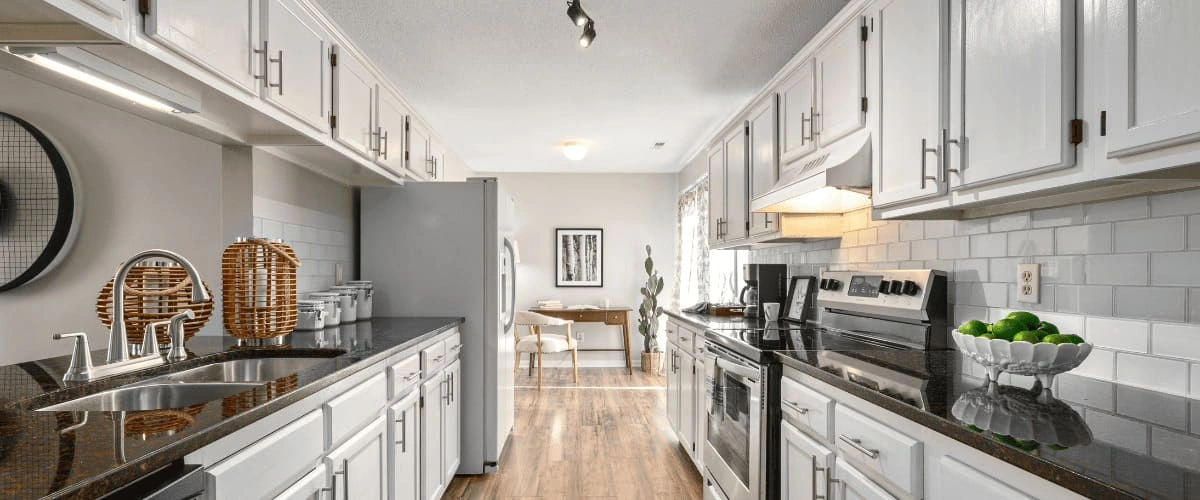 A long galley-style kitchen featuring bright white cabinetry, dark granite countertops, a classic white subway tile backsplash, and stainless steel appliances.