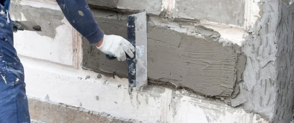 A worker in blue overalls applying a thick layer of grey cement mortar to a foundation wall using a large metal hand trowel for waterproofing and smoothing.