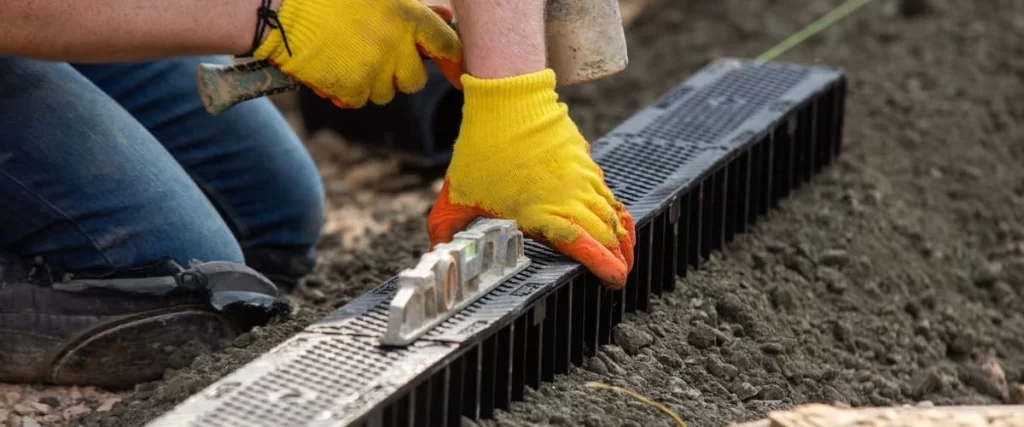 A contractor wearing yellow gloves uses a spirit level to align a black plastic drainage channel in the soil next to a building foundation.