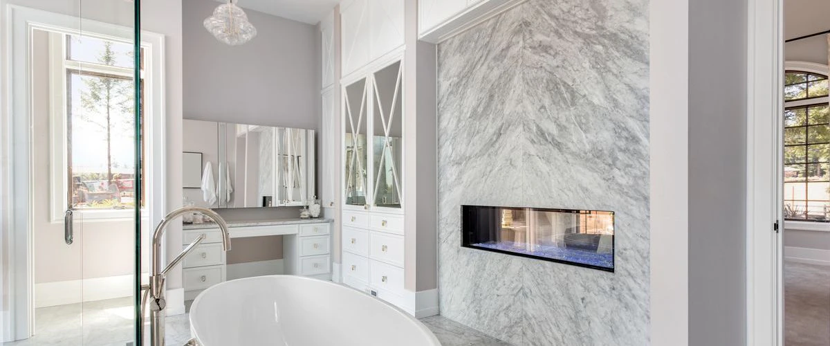 A high-end luxury bathroom featuring a white freestanding soaking tub positioned in front of a book-matched marble accent wall with a built-in fireplace.