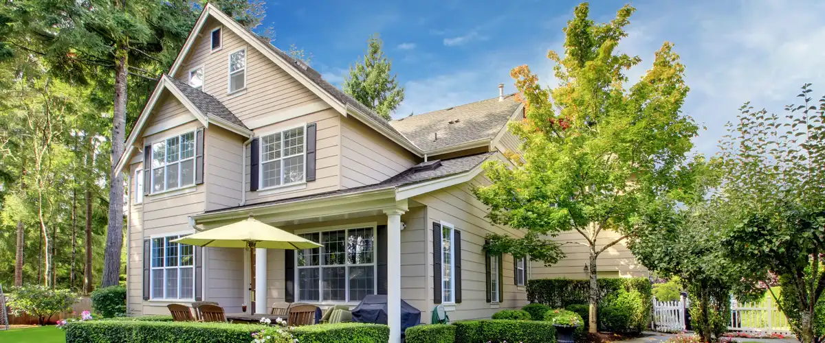 Side view of a spacious beige two-story home with dark shutters, featuring an outdoor patio area with a yellow umbrella, dining set, and lush green hedges.