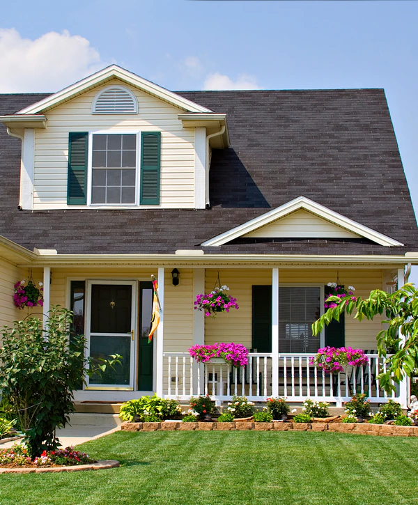 A charming yellow two-story house in Buena Park, CA, constructed by a professional home builder, featuring a black shingle roof and white porch railings with purple flowers.