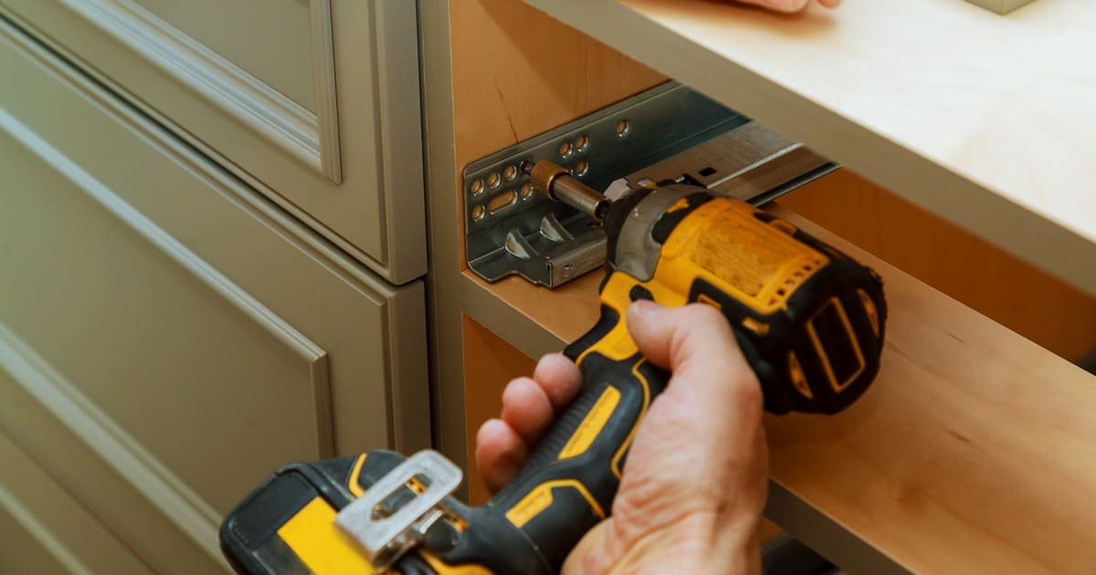 A close-up of a worker using a yellow cordless drill to install metal drawer slides inside a wooden cabinet, demonstrating tasks that factor into handyman jobs cost.
