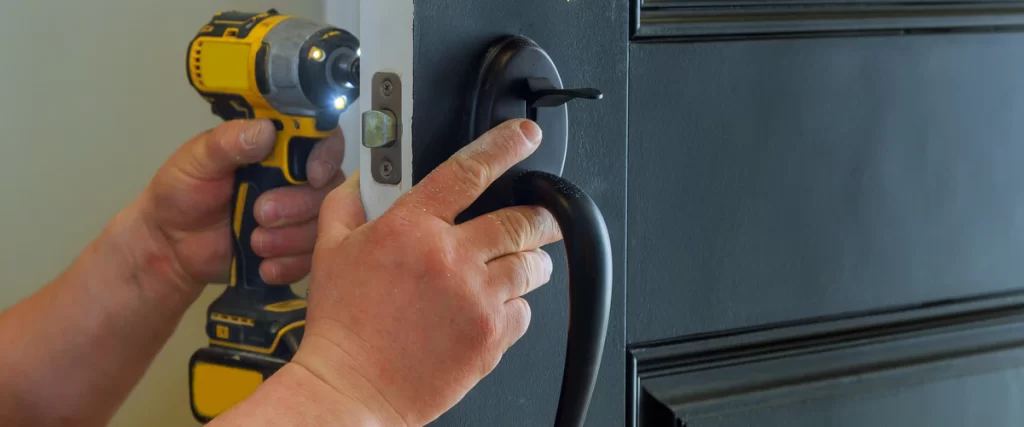 A close-up view of a handyman using a yellow cordless drill to carefully install a black lock and handle onto a dark-colored door.