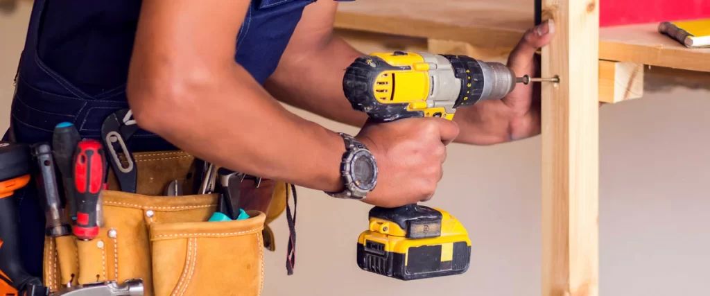 A professional handyman wearing a loaded leather tool belt uses a yellow cordless drill to secure a screw into a wooden frame structure.