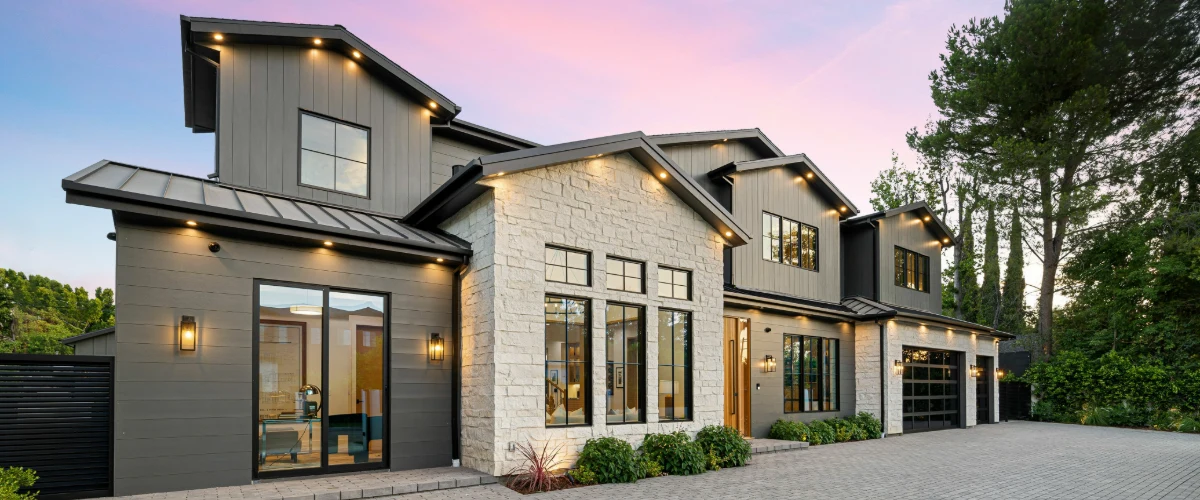 A large, modern home exterior featuring grey siding, white stone accents, black window frames, and warm exterior lighting at dusk.